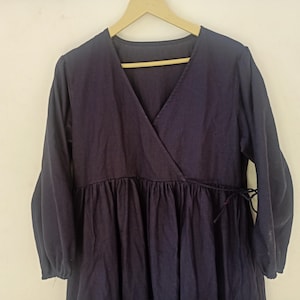 Puede incluir: Un vestido cruzado azul marino con mangas largas y cintura fruncida. El vestido tiene un escote en V y está colgado de una percha de madera. La tela parece ser un material suave y tejido.