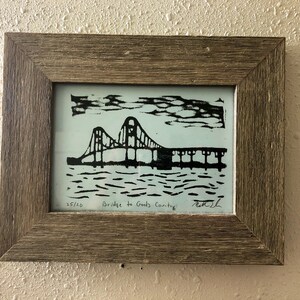 Puede incluir: Una impresión en blanco y negro enmarcada de un puente con el texto "Bridge to Gods Country" y el número "25/20" en la esquina inferior izquierda. La impresión está firmada "Matt L" en la esquina inferior derecha.