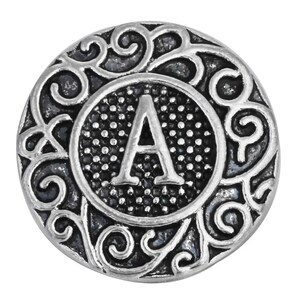 May include: A silver-colored, round button with the letter "A" in the center. The letter is inside a dotted circle, surrounded by an ornate, swirling design. The button has a vintage or antique aesthetic.
