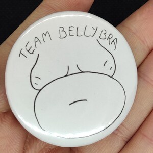 Könnte beinhalten: Runder weißer Knopf mit einer schwarzen Linienzeichnung eines Bauches mit dem Text "TEAM BELLY BRA" oben.