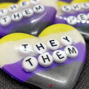 May include: Heart-shaped resin art pieces in the non-binary pride flag colors: yellow, white, purple, and gray. Each heart has the words "THEY THEM" spelled out in white letter beads. The background is a dark gray.