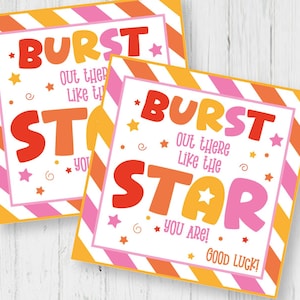 Puede incluir: Dos etiquetas cuadradas con el texto "BURST OUT THERE LIKE THE STAR YOU ARE! GOOD LUCK!" en rosa, naranja y blanco. Las etiquetas miden 5,8 x 5,8 cm. También se ven una hoja de etiquetas y unas tijeras.