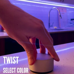 Op de afbeelding: Een ronde witte knop met de woorden "TWIST SELECT COLOR" gedrukt op een wit oppervlak.
