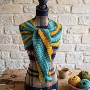 Peut inclure: Un châle tricoté présentant des rayures diagonales dans des tons de bleu sarcelle, jaune et bleu foncé, drapé sur un torse de mannequin recouvert d'un tissu à carreaux. Une petite étiquette rectangulaire avec le texte manuscrit Breizhlaine est fixée.