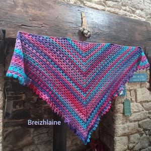 Peut inclure: Un châle crocheté fait à la main, aux couleurs vives de rose, violet et turquoise. Le châle est drapé sur une poutre en bois. Le texte "Breizhaine" est visible en bas à gauche. Le châle a un bord festonné.