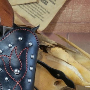 Könnte beinhalten: Ein schwarzes Lederholster mit roten Nähten und silbernen Nieten. Das Holster ist für eine Pistole ausgelegt und hat ein dekoratives Blumenmuster. Das Holster liegt auf einer Holzoberfläche mit einem Paar Sporen und einer Zeitung.