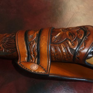 May include: A brown leather holster with floral and leaf carvings, designed to fit a revolver. The holster features a decorative copper rivet and a stitched border. The leather has a rich, warm tone.