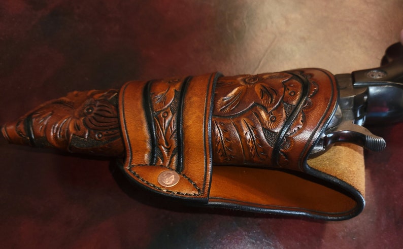 May include: A brown leather holster with floral and leaf carvings, designed to fit a revolver. The holster features a decorative copper rivet and a stitched border. The leather has a rich, warm tone.