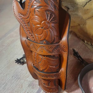 May include: A brown leather holster with intricate floral carvings, designed to hold a pistol. The holster is decorated with a repeating pattern of small, decorative elements.
