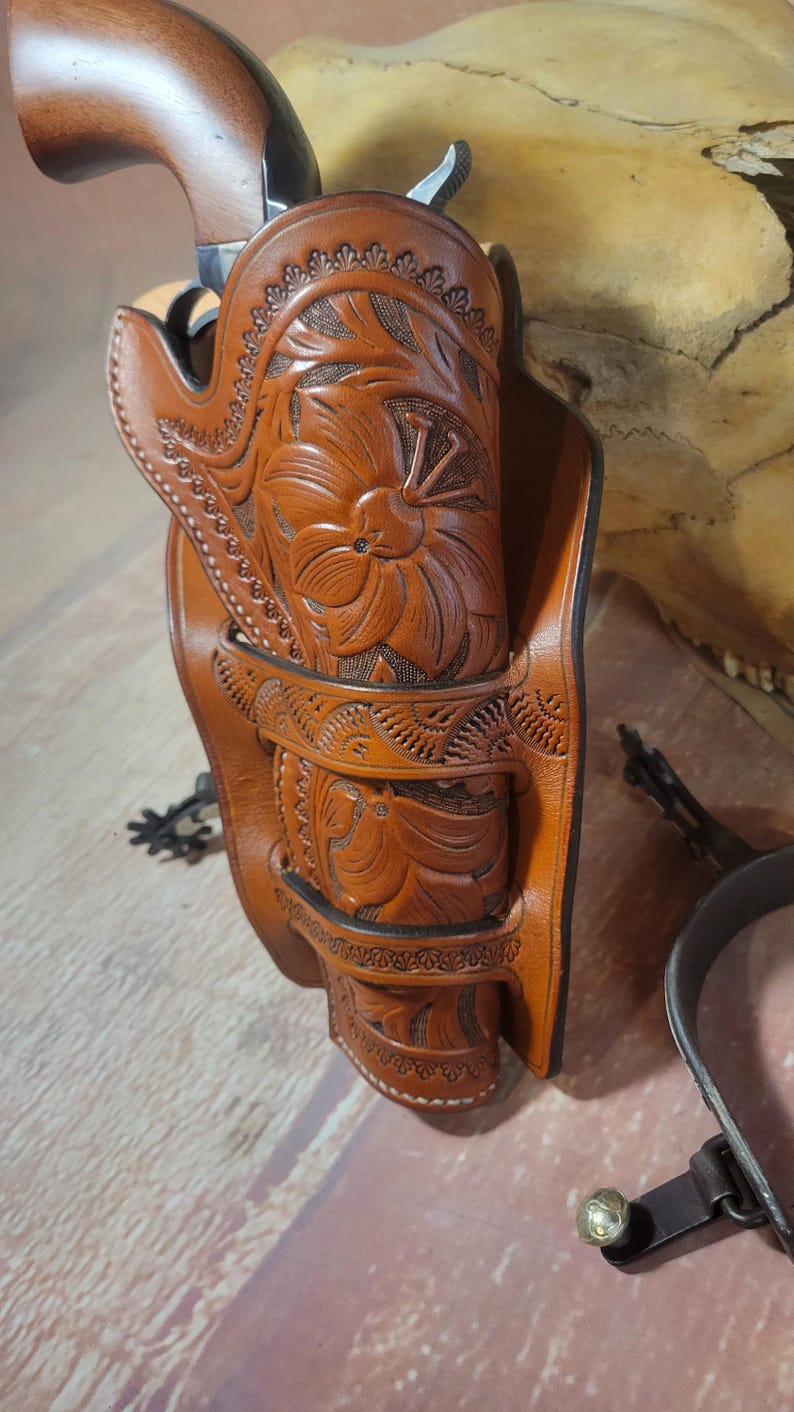 May include: A brown leather holster with intricate floral carvings, designed to hold a pistol. The holster is decorated with a repeating pattern of small, decorative elements.