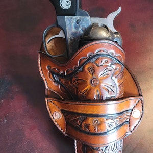 May include: A vintage revolver in a brown leather holster with floral tooling. The holster has a detailed carved floral design and is secured with metal rivets. The text "Floral A" is visible.