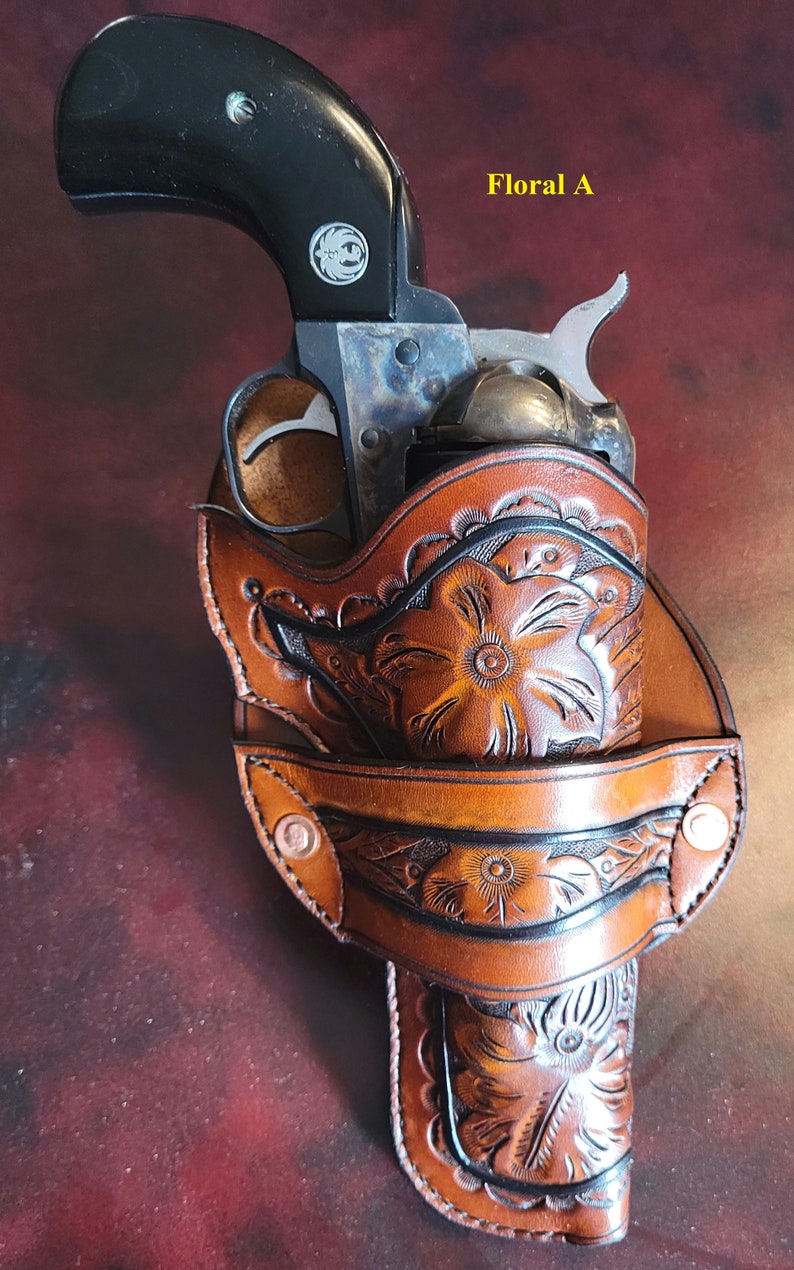 May include: A vintage revolver in a brown leather holster with floral tooling. The holster has a detailed carved floral design and is secured with metal rivets. The text "Floral A" is visible.