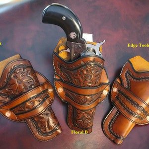May include: Three brown leather gun holsters with floral and edge-tooled designs. One holster holds a black-handled revolver. The holsters are displayed on a dark brown surface. The text "Floral A", "Floral B", and "Edge Tooled" are visible.