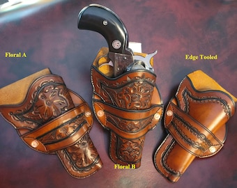 May include: Three brown leather gun holsters with floral and edge-tooled designs. One holster holds a black-handled revolver. The holsters are displayed on a dark brown surface. The text "Floral A", "Floral B", and "Edge Tooled" are visible.