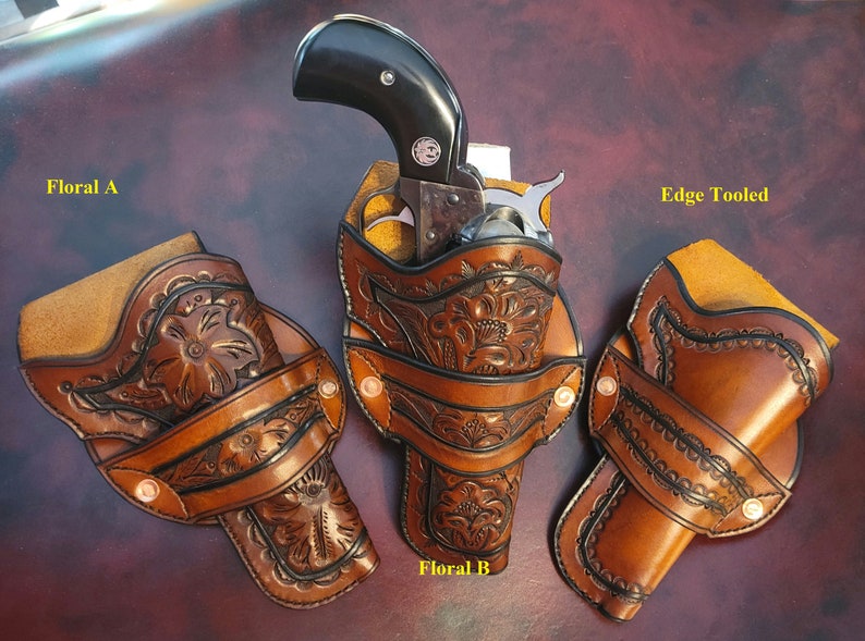May include: Three brown leather gun holsters with floral and edge-tooled designs. One holster holds a black-handled revolver. The holsters are displayed on a dark brown surface. The text "Floral A", "Floral B", and "Edge Tooled" are visible.