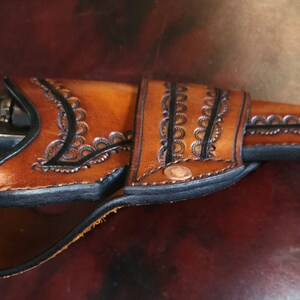 May include: A handcrafted brown leather holster with intricate floral tooling and black edging. The holster is designed to fit a revolver, with a copper rivet detail. The leather has a rich, warm tone, suggesting a vintage or Western aesthetic.