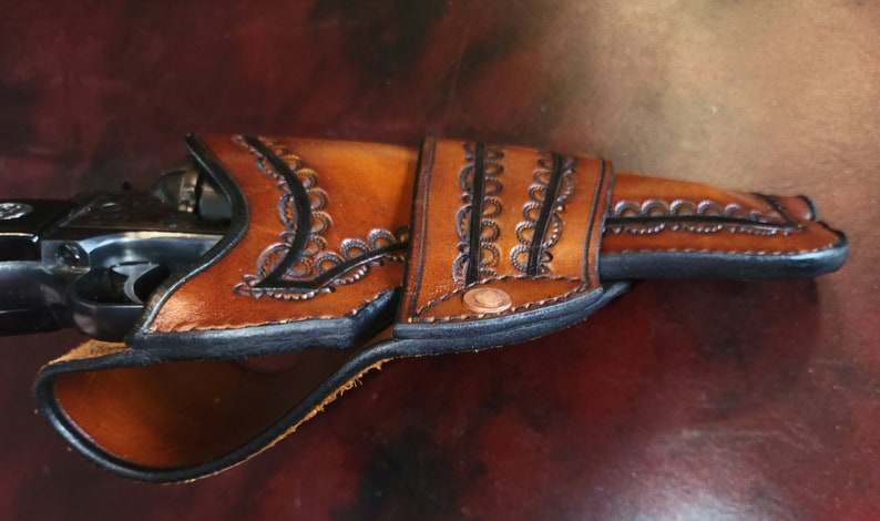 May include: A handcrafted brown leather holster with intricate floral tooling and black edging. The holster is designed to fit a revolver, with a copper rivet detail. The leather has a rich, warm tone, suggesting a vintage or Western aesthetic.