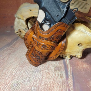 May include: A brown leather holster with intricate floral designs, holding a black handgun. The holster is resting on a light brown wooden surface.