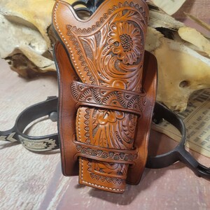 May include: A brown leather holster with intricate floral tooling, designed to hold a handgun. The holster is displayed on a wooden surface with a newspaper article about the Younger Gang.