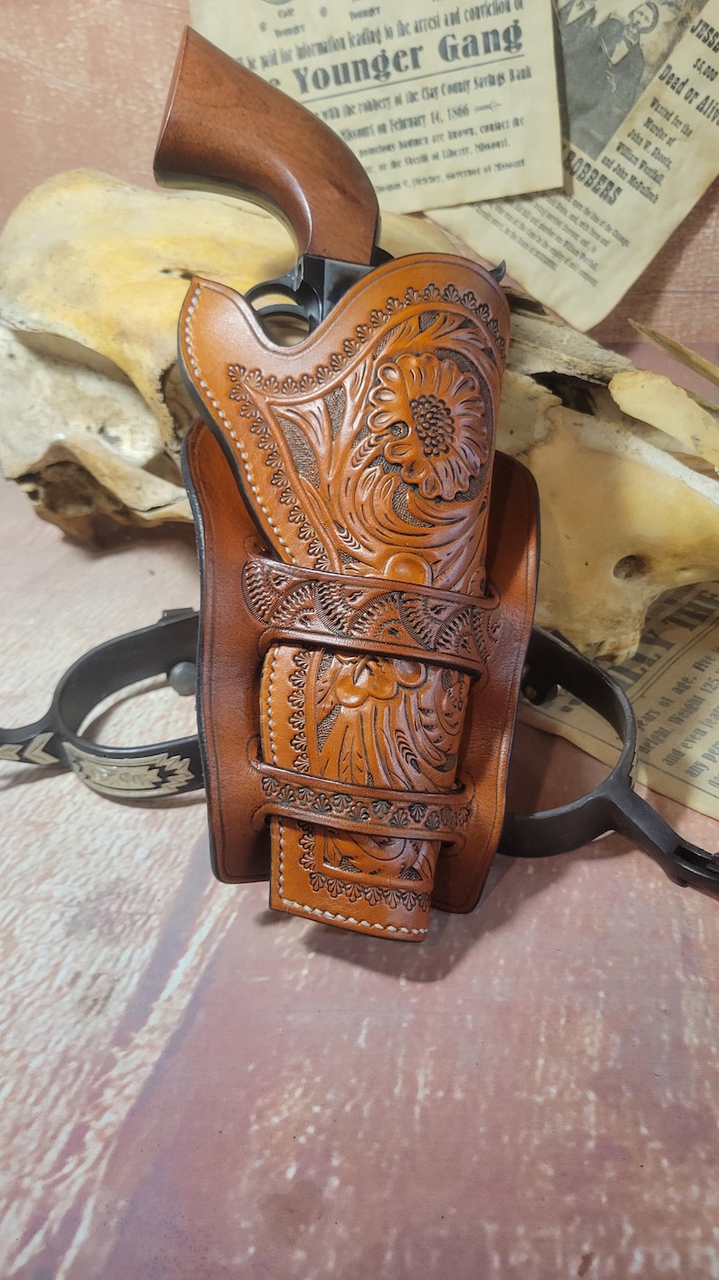 May include: A brown leather holster with intricate floral tooling, designed to hold a handgun. The holster is displayed on a wooden surface with a newspaper article about the Younger Gang.