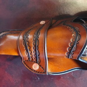 May include: A handcrafted brown leather holster with decorative black edging and copper rivets. The holster features intricate floral-like cutouts and holds a silver and black revolver. The leather has a rich, warm tone.