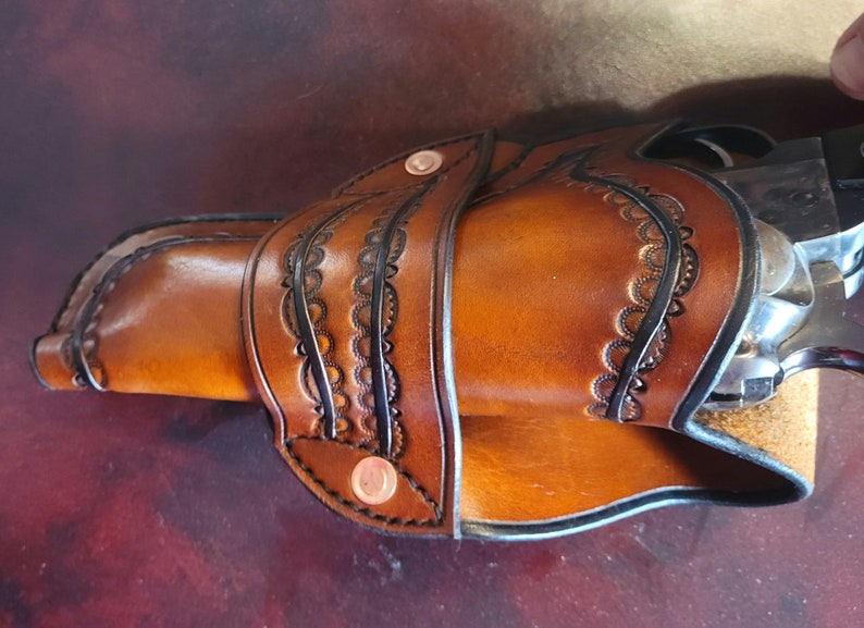 May include: A handcrafted brown leather holster with decorative black edging and copper rivets. The holster features intricate floral-like cutouts and holds a silver and black revolver. The leather has a rich, warm tone.