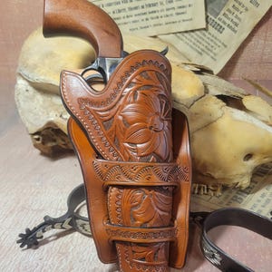 May include: A brown leather holster with intricate floral carvings, designed to hold a revolver. The holster has a belt loop and is decorated with stitching. The revolver is partially visible in the holster.