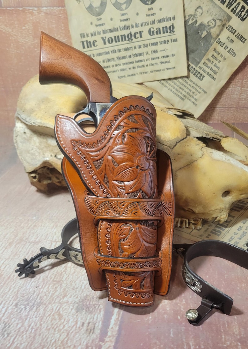 May include: A brown leather holster with intricate floral carvings, designed to hold a revolver. The holster has a belt loop and is decorated with stitching. The revolver is partially visible in the holster.