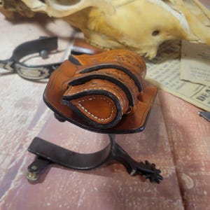 May include: A brown leather holster with intricate stitching and a decorative design. The holster is designed to hold two small cylindrical objects, possibly bullets or cartridges.