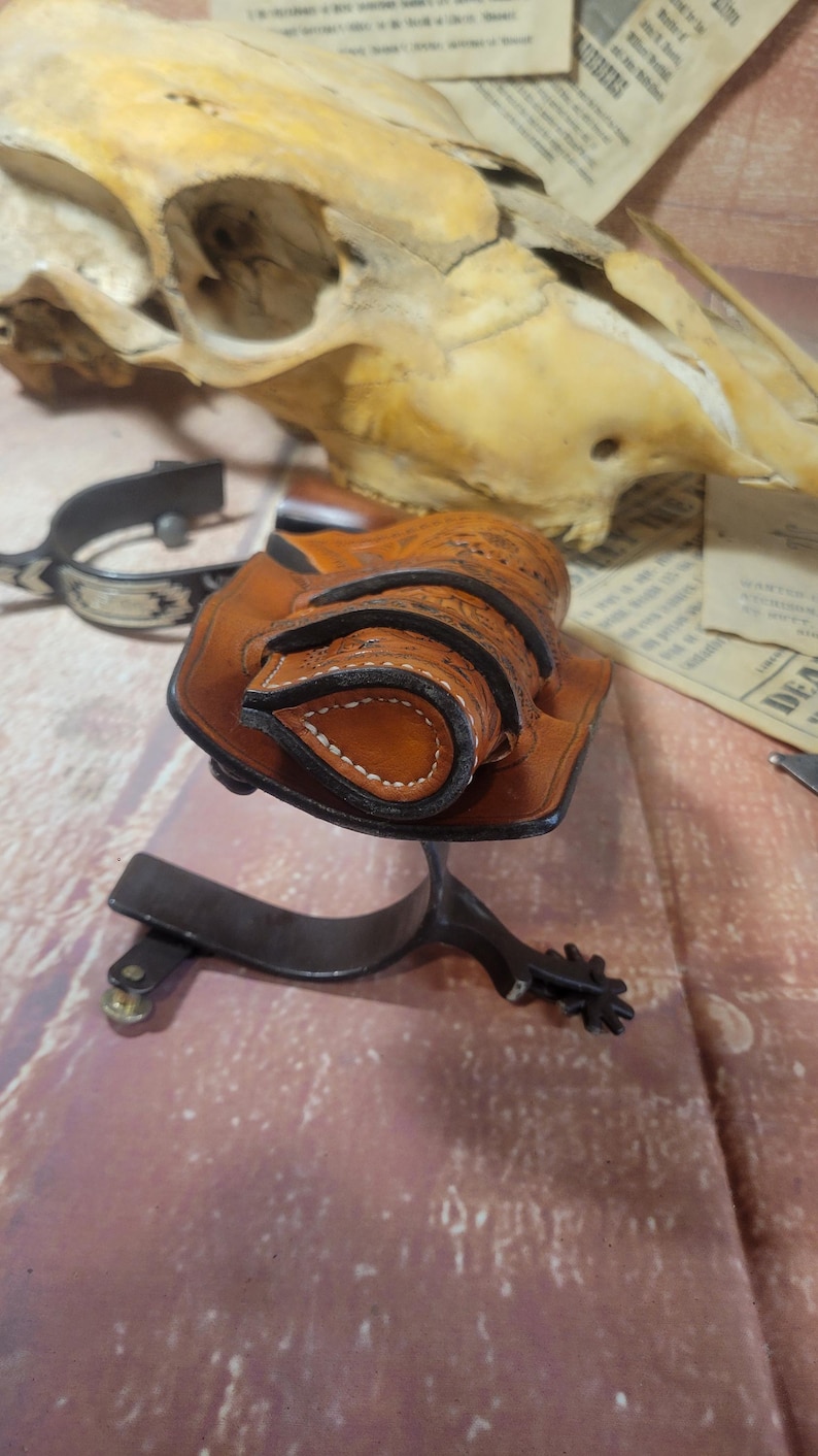 May include: A brown leather holster with intricate stitching and a decorative design. The holster is designed to hold two small cylindrical objects, possibly bullets or cartridges.