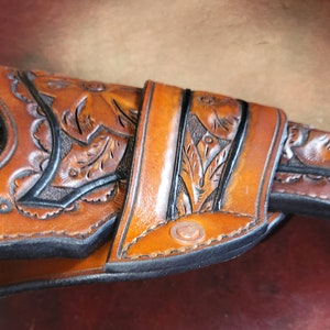 May include: A brown leather holster with floral tooling, holding a revolver. The holster features intricate leaf and vine designs, with a dark border. The leather has a rich, warm tone, and the revolver is partially visible.