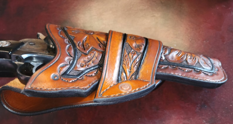 May include: A brown leather holster with floral tooling, holding a revolver. The holster features intricate leaf and vine designs, with a dark border. The leather has a rich, warm tone, and the revolver is partially visible.