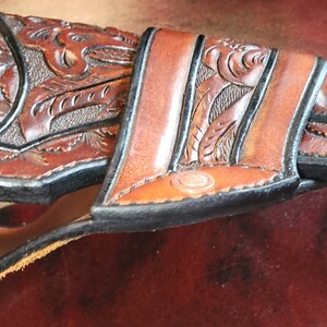 May include: A brown leather holster with intricate floral and leaf tooling. The holster is designed to fit a revolver, with a detailed carved design and a contrasting black edge. The leather has a rich, warm tone, and the craftsmanship is evident in the fine details.