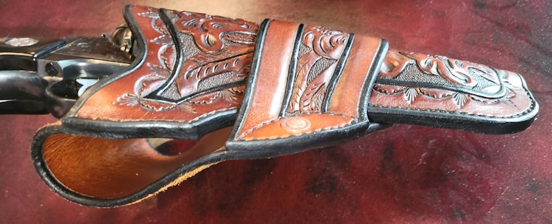 May include: A brown leather holster with intricate floral and leaf tooling. The holster is designed to fit a revolver, with a detailed carved design and a contrasting black edge. The leather has a rich, warm tone, and the craftsmanship is evident in the fine details.