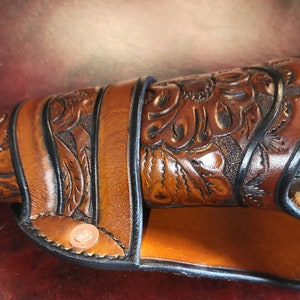 May include: A brown leather holster with intricate floral tooling, holding a silver revolver. The holster features a decorative design and is secured with a strap and button closure. The revolver's details are visible.