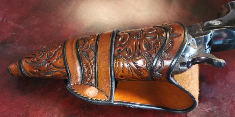 May include: A brown leather holster with intricate floral tooling, holding a silver revolver. The holster features a decorative design and is secured with a strap and button closure. The revolver's details are visible.