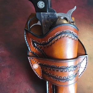 May include: A brown leather holster with a decorative edge-tooled design, holding a black-handled revolver. The holster features a detailed pattern and is secured with rivets. The text "Edge Tooled" is visible.