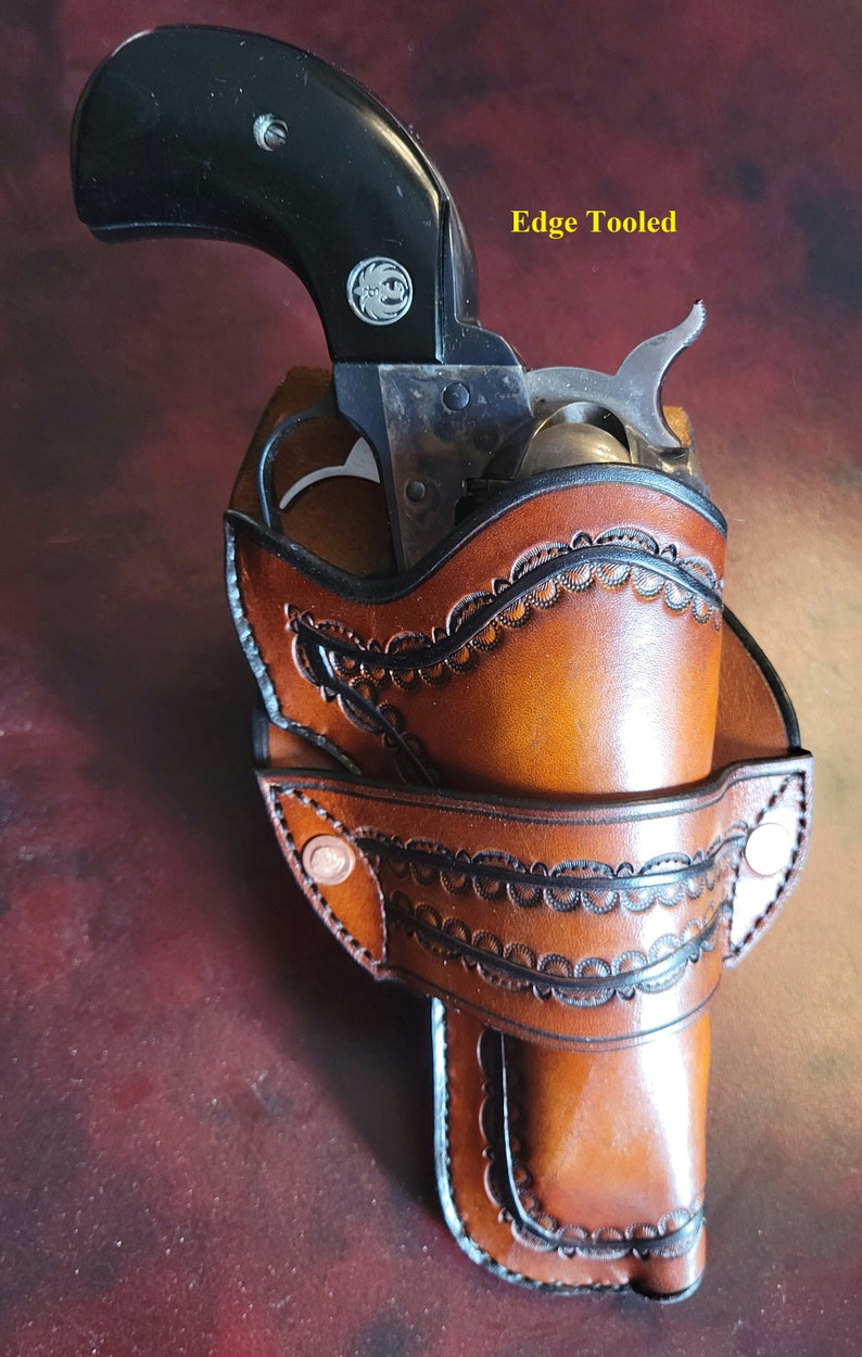 May include: A brown leather holster with a decorative edge-tooled design, holding a black-handled revolver. The holster features a detailed pattern and is secured with rivets. The text "Edge Tooled" is visible.