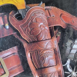 May include: A close-up of a brown leather holster with intricate floral carvings. The holster is designed to hold a pistol and has a metal buckle. The text "PACKING IRON" and "LEATHER OF THE FRONTIER WEST" is visible at the top of the image. The text "RICHARD C. RATTENBURY" is visible at the bottom of the image.