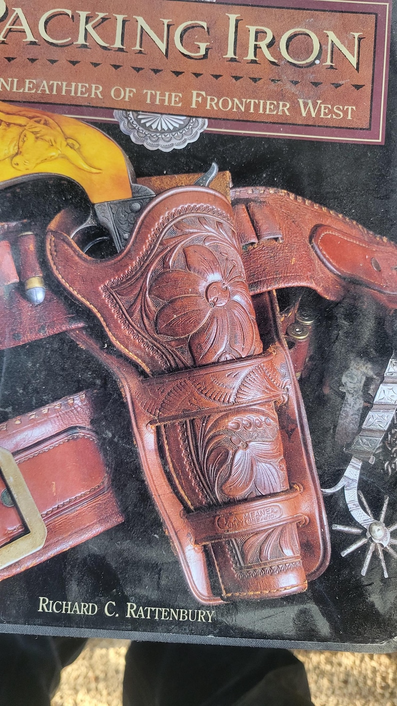 May include: A close-up of a brown leather holster with intricate floral carvings. The holster is designed to hold a pistol and has a metal buckle. The text "PACKING IRON" and "LEATHER OF THE FRONTIER WEST" is visible at the top of the image. The text "RICHARD C. RATTENBURY" is visible at the bottom of the image.