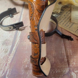 May include: A brown leather holster with intricate floral carvings. The holster is designed to hold a handgun and has a belt loop.