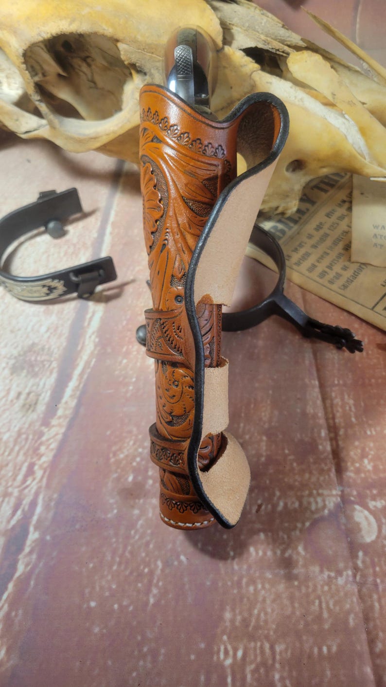 May include: A brown leather holster with intricate floral carvings. The holster is designed to hold a handgun and has a belt loop.