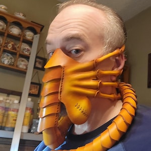 May include: A handcrafted, orange leather mask with a segmented design, featuring a detailed, textured appearance. The mask has a distinctive, stylized shape with a neck piece, and is likely a costume or cosplay item.