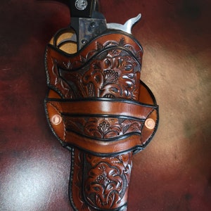 May include: A brown leather holster with intricate floral tooling. The holster is designed to hold a revolver, with a black handle visible. The text "Floral B" is in the top right corner.