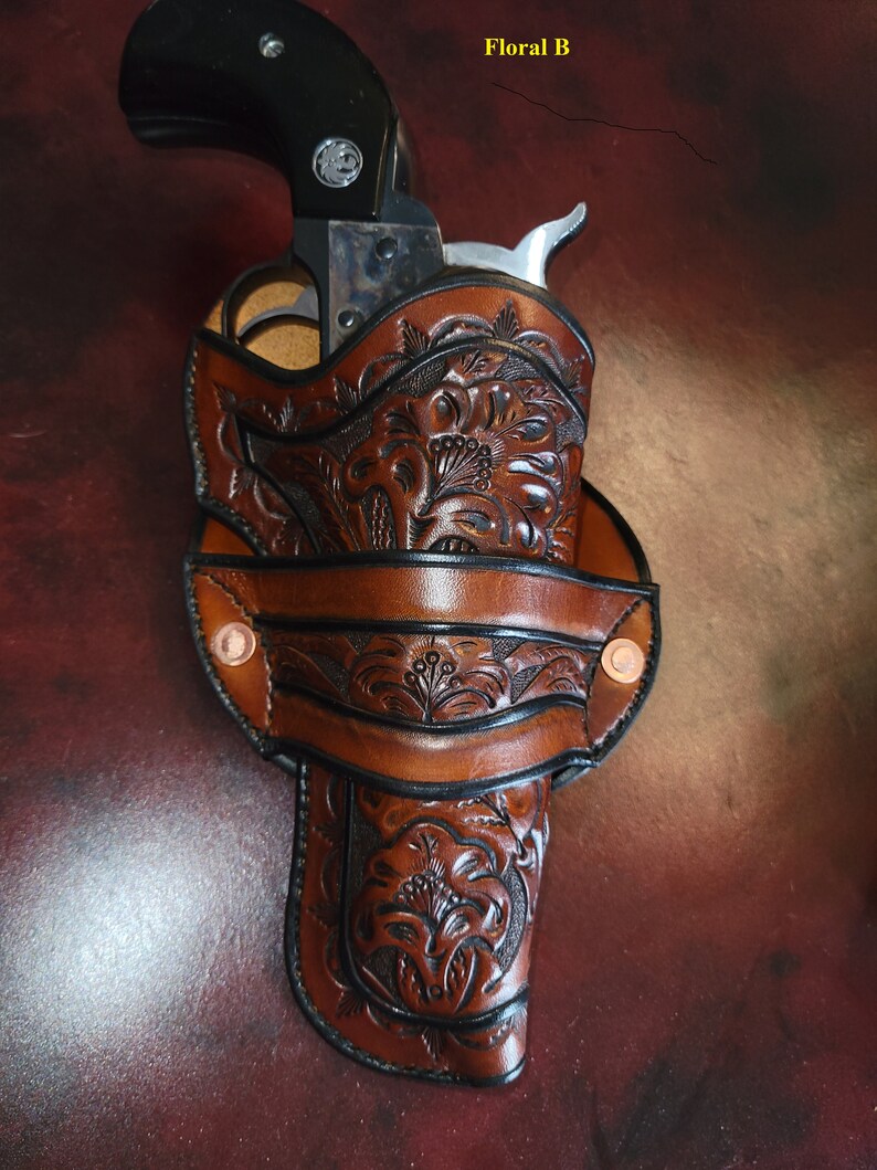May include: A brown leather holster with intricate floral tooling. The holster is designed to hold a revolver, with a black handle visible. The text "Floral B" is in the top right corner.