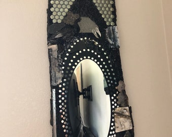 May include: A rectangular wall mirror with an oval mirror in the center. The mirror is framed with black mosaic tiles, stones, and decorative elements. The top section features a pattern of small, round, silver tiles. The mirror reflects a room.