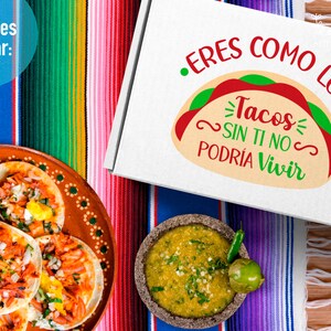 May include: A white box with the text "Eres como los Tacos Sin Ti No Podria Vivir" in green and red lettering. The box is sitting on a colourful striped blanket with a plate of tacos and a bowl of salsa.