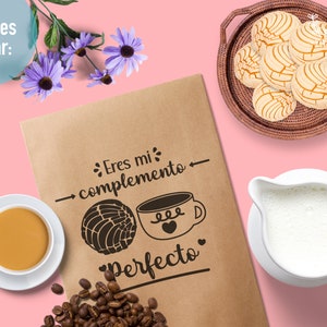 May include: A brown paper bag with the text "Eres mi complemento Perfecto" and a drawing of a coffee cup and a pastry. The bag is on a pink background with a cup of coffee, a jug of milk, and a plate of pastries.