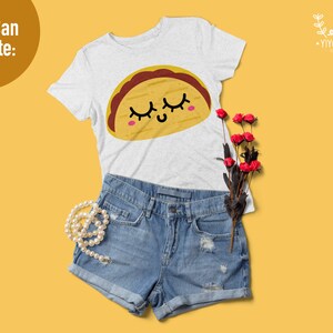K&ouml;nnte beinhalten: Ein wei&szlig;es T-Shirt mit einem Cartoon-Taco-Design. Der Taco hat ein l&auml;chelndes Gesicht mit geschlossenen Augen und Wimpern. Das Shirt wird mit Jeans-Shorts und einer Perlenkette kombiniert.