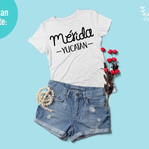 May include: A white t-shirt with the text "M&eacute;rida -YUCAT&Aacute;N- " in black lettering. The shirt is laid out on a light blue background with a pair of blue denim shorts, a pearl necklace, and a bouquet of red roses.